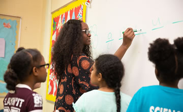 a photo of a student teacher writing on a white board with young students gathered around her